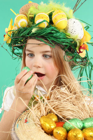 Cute little child wearing a fun hairstyle on Easter Day. Girl with basket of shiny eggs looks at an easter egg squintingの写真素材