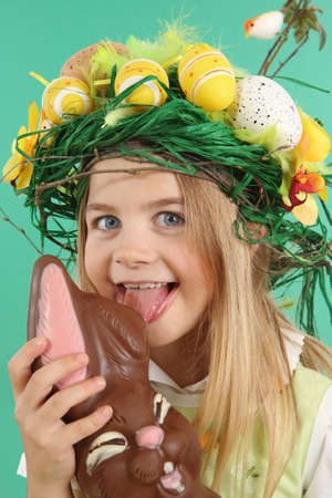 Easter concept. Child eating chocolate bunny. The young Girl wears a festive headdress made of yellow Easter eggs, spring flowers and bird feathers.の写真素材