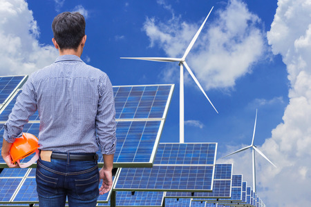 engineer stand holding yellow construction helmet in solar farm and wind turbines generating...