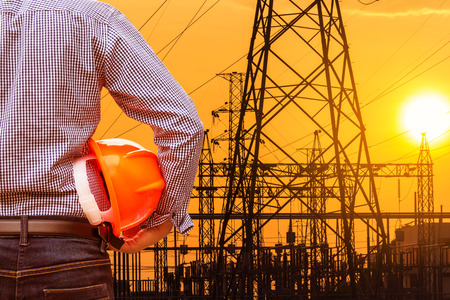 engineer holding yellow safety helmet with high voltage electric pylon substation on sunset