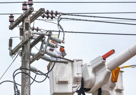 Electrician in bucket hydraulic lifting platform while repairing wire of the power line on electricity power pole with blue skyの写真素材