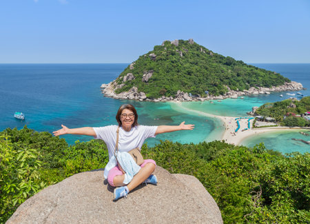 Woman sitting on stone high cliff raising hands open arms achievements celebrate enjoy after climbing to the peak of the hill  at Koh Nang Yuan Island ,Surat Thani, Thailandの写真素材