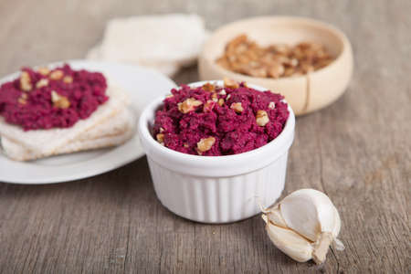 Beetroot pesto in a white bowl on a wooden table with garlic beetroot and rice cakes on the back,...