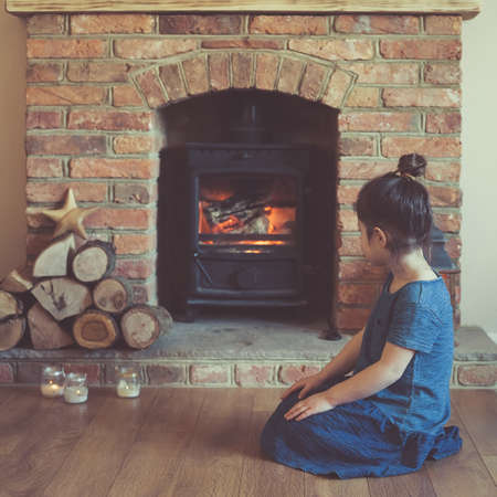 Little girl sitting in front of the fireplace looking at the fire flame in the woodburner, dark toned photo, selective focus, squareの写真素材