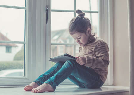 Toned photo of beautiful little girl sitting on the window holding the tablet, selective focusの写真素材