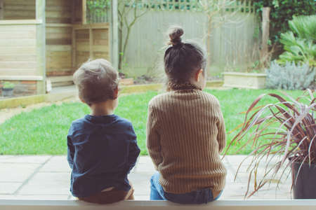Two little girls sitting on the floor indoors looking at the garden; opened patio doors, selective focusの写真素材