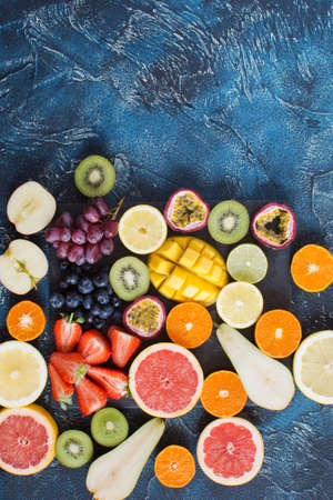 Fruits in rainbow colors, oranges, lemons, kiwis, grapefruits, strawberries, grapes, blueberries, pears, passion fruit, mango on the black slate on the dark blue background, top viewの写真素材