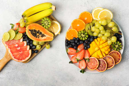 Healthy fruits and berries in rainbow colours on a white plate and wooden board, strawberries, mango, grapes, bananas, grapefruit on the light grey table, copy space, top view, selective focusの写真素材