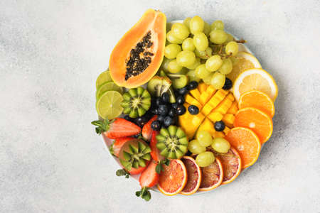 Healthy fruits and berries in rainbow colours on a white plate, strawberries, mango, grapes, bananas, grapefruit on the light grey table, copy space for text, top view, selective focusの写真素材