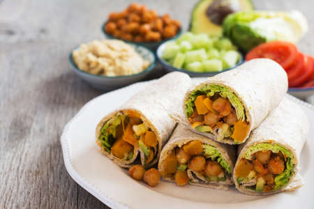 Delicious vegatarian wraps made with cajun chickpeas, butternut squash, spring onion, cucumber, avocado with ingredients on the back on the dark wooden table, copy space for textの写真素材