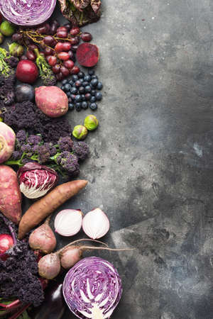 Vertical photo, vegetables and fruits in purple, green and dark red colours, cabbage kale sprouts sweet poato onions broccoli blueberries grapes plums, top view, grey background, selective focusの写真素材