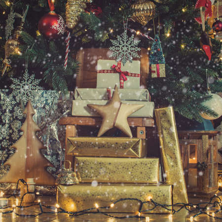 Stack of presents under lit up Christmas tree with baubles and ornaments, lantern, stars and garlands, created snow, selective focus, tonedの写真素材