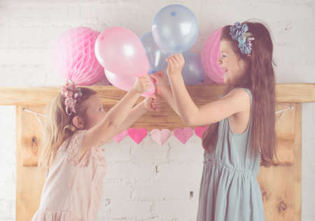 Two little girls in pastel pink and blue dresses, floral headbands holding pastel colored balloons,...