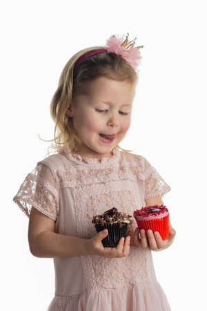 Little girl in pastel pink dress and crown headband holding chocolate cupcake and red cupcake, licking her lips, on white, selective focusの写真素材