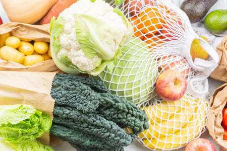 Assortment of vegetables fruits in reusable white mesh, cotton and paper bags on white kitchen...