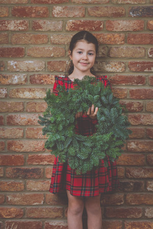 Little girl in red tartan dress holding green handmade Christmas wreath, brick wall background, selective focus, tonedの写真素材