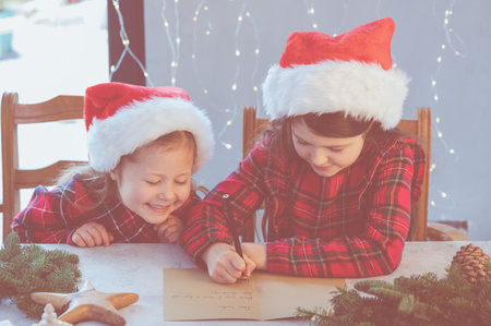 Little girls in red Chirstmas hats and red tartan dresses writing letter to Santa Claus sitting at the table indoors, beautiful light bokeh, selective focus.の写真素材