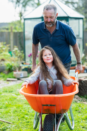 Father riding the wheelbarrow with a little girl in it in the garden. Smiley girl with a long hair in the orange barrow, selective focusの写真素材