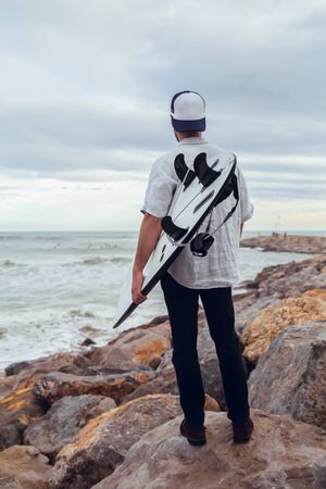 Boy from behind with surfboard cap and shirt on rocky beach watching the sea in Barcelonaの写真素材
