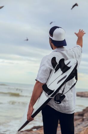 Boy from behind with surfboard cap and shirt raising hand with birds in the background on rocky beach looking at the sea in Barcelonaの写真素材