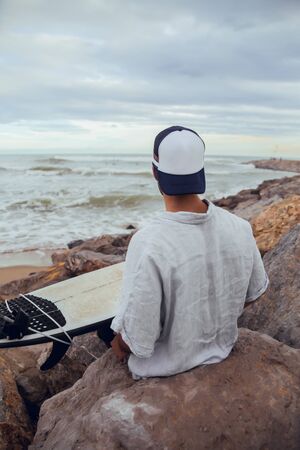 Boy from behind from beach with cap and white surfboard looking at the sea with cloudy dayの写真素材