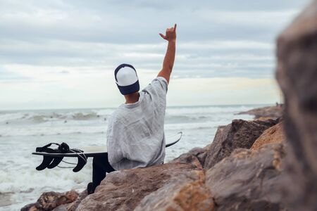 Boy with his back raised from beach with cap and white surfboard looking at the sea with cloudy dayの写真素材