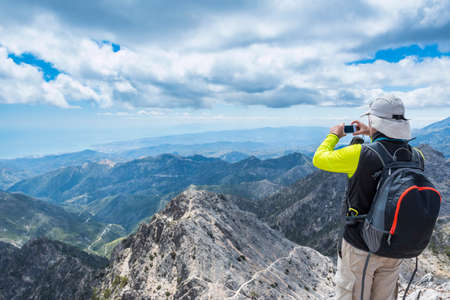 Back view of a hiker standing at the edge and taking shots of the mountainscapeの写真素材