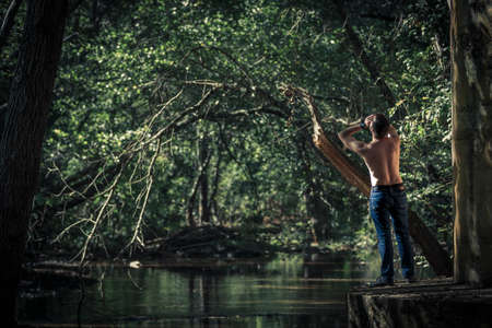 Back view of shirtless man in jeans standing and shouting on background of green pond and treesの写真素材