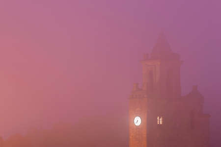 View of old stone bell tower with clock of a church standing in thick fogの写真素材
