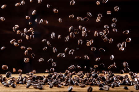 Dark-brown roasted coffee beans in freeze flying in air above wooden tablet against black backgroundの写真素材