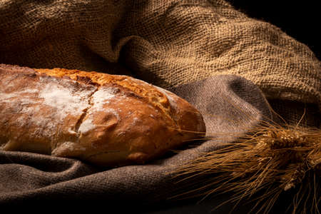 Big brown baked loaf of bread on canvas material with wheat on dark background