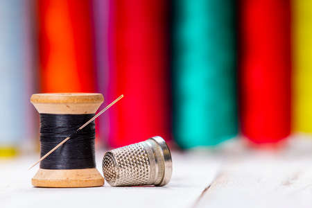 Close-up view of wooden sewing spool with black thread and needle and shiny metal thimble with assorted buttons placed on white table on blurred backgroundの写真素材