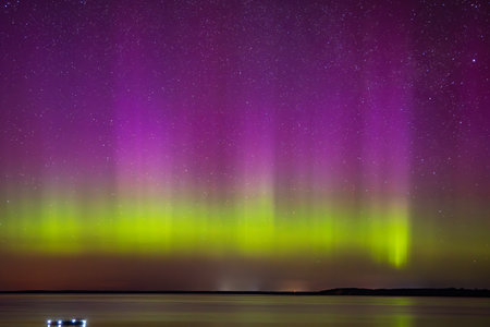 Colorful northern lights with vivid purple and green hues dance across a star-filled night sky above a calm lake horizon.の写真素材