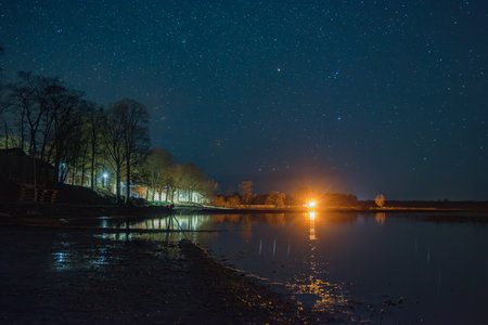 Starry night sky over a quiet lakeside landscape with sparkling lights and a camera on the horizon,...