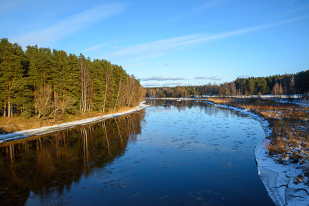 A partially frozen river winding through a winter landscape, surrounded by trees and snow-covered...