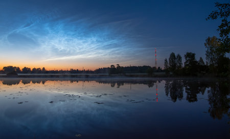 Twilight view of a calm lake with reeds, pier stubs, noctilucent clouds, and a red-lit radio tower reflected in the water near forest edge,silver cloudsの写真素材