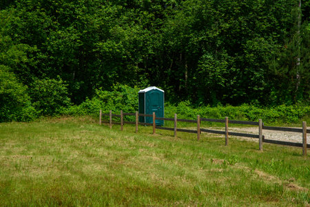 A lone portable toilet stands by a wooden fence in a grassy field, with dense green forest backdrop on a sunny summer dayの写真素材