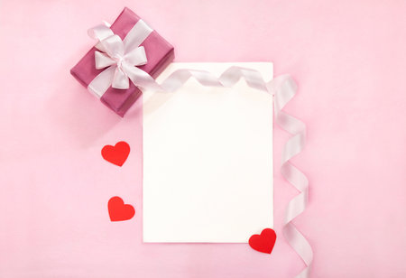 Valentines day greeting card with pink gift box, white bow, long curved ribbon and paper red hearts. Tope view, place for textの写真素材