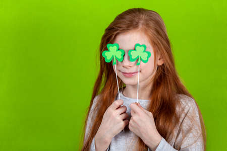 Portrait of little smiling caucasian red-haired girl covering her eyes with shamrock clover leaves on sticks on green studio background.. Irish St. Patricks Day. Copyspace.の写真素材