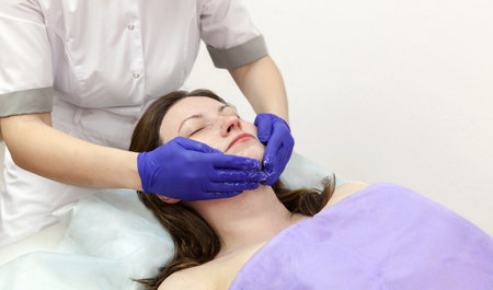 Cosmetologist hands in blue protective gloves touch face of caucasian woman patient making massage procedure in beauty salon. Concept of rejuvenation, skin care and facial treatment.の写真素材