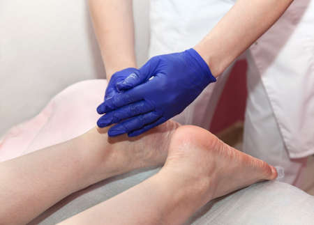 Masseur hands in blue protective gloves touch heels of woman patient doing foot massage procedure in...
