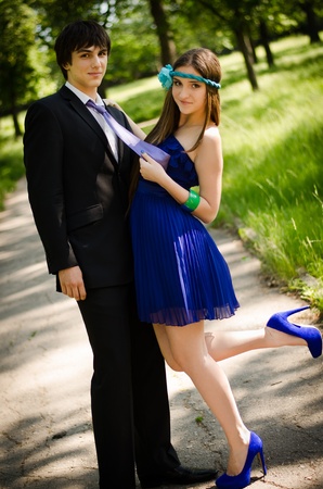 Young couple of high school seniors after the prom in a summer park. Beautiful brunette in blue dress and shoes pulling man's tie.の写真素材