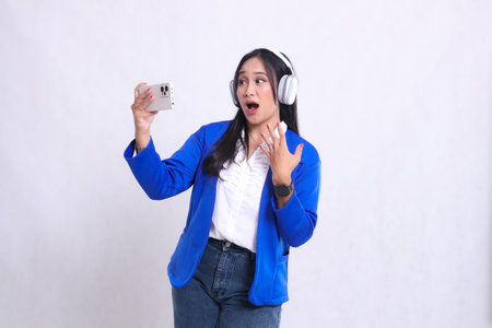 beautiful mature office woman wearing blue formal suit standing on right side shocked losing playing on smartphone mobile phone and headphone isolation white background. for entertainment contentの写真素材