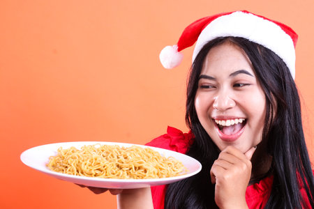 close up of cheerful young indonesia woman eating noodles, hands holding white plate filled with noodles to the side and chin, wearing red Christmas blouse, Santa hat, isolated on orange backgroundの写真素材