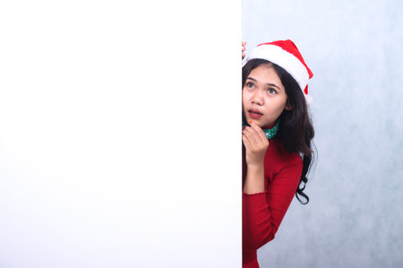 expression of adult Asian woman wearing christmas sweater, Santa hat and scarf, imagining thinking to the right up holding chin, isolated behind blank white bannerの写真素材