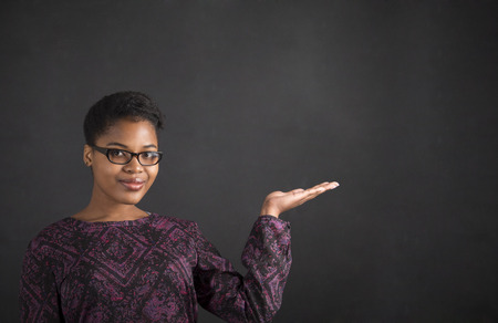 South African or African American black woman teacher or student holding her hand out to the side standing against a chalk blackboard background insideの写真素材