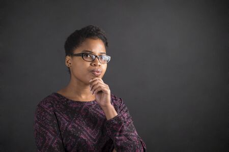 South African or African American black woman teacher or student with her hand on her chin whilst thinking standing against a chalk blackboard background insideの写真素材