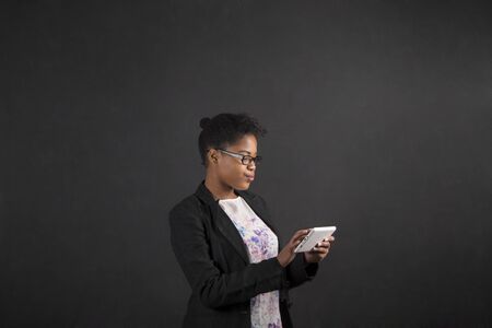 South African or African American black woman teacher or student holding a tablet standing against a chalk blackboard background insideの写真素材