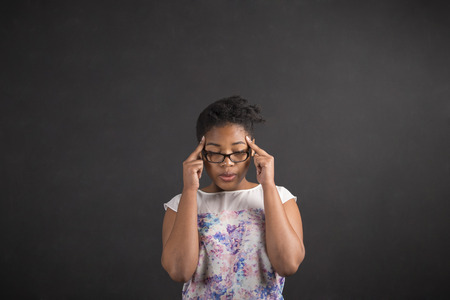 South African or African American black woman teacher or student holding her fingers on her temples thinking against a chalk black board background insideの写真素材