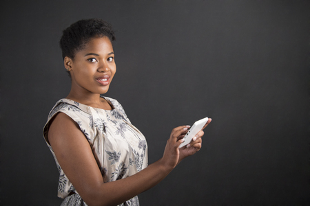 South African or African American black woman teacher or student holding a tablet standing against a chalk blackboard background insideの写真素材